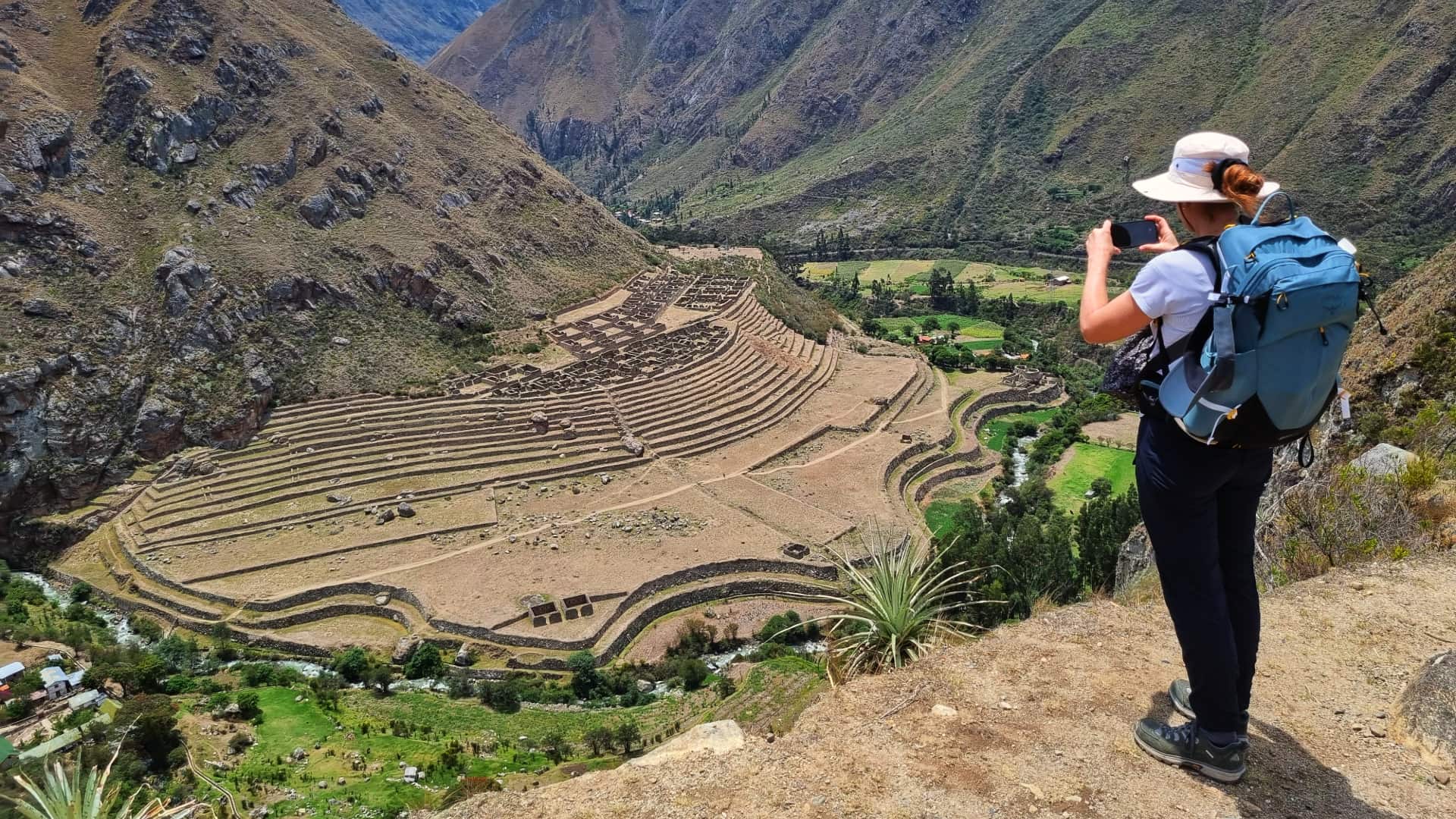 A tourist holding a camera, photographing the picturesque Sacred Valley, with terraced fields, lush greenery, and towering mountains in the background. A tourist holding a camera, photographing the picturesque Sacred Valley, with terraced fields, lush greenery, and towering mountains in the background.