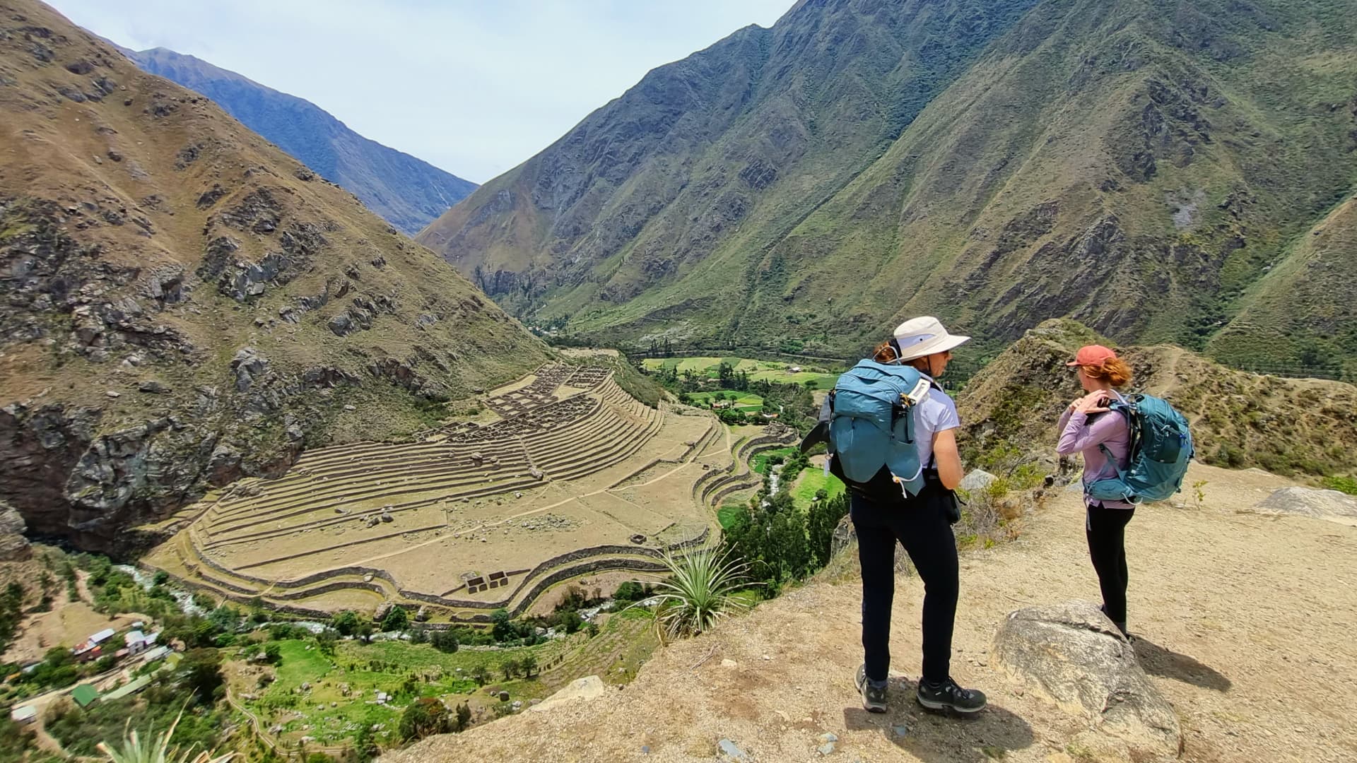 Two tourists standing on a viewpoint overlooking the Sacred Valley, gazing at the terraced fields and towering mountains under a clear blue sky. Two tourists standing on a viewpoint overlooking the Sacred Valley, gazing at the terraced fields and towering mountains under a clear blue sky.