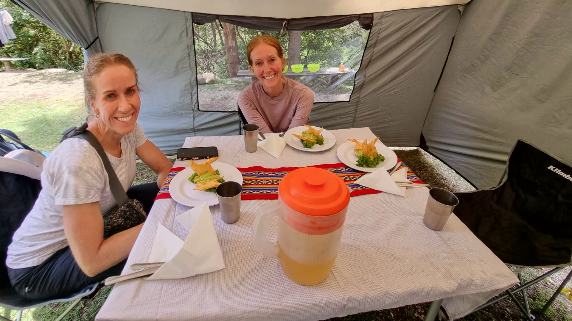 Two women sitting inside a camping tent, enjoying a warm breakfast with scenic Andean mountains visible through the tent's open door. Two women sitting inside a camping tent, enjoying a warm breakfast with scenic Andean mountains visible through the tent's open door.