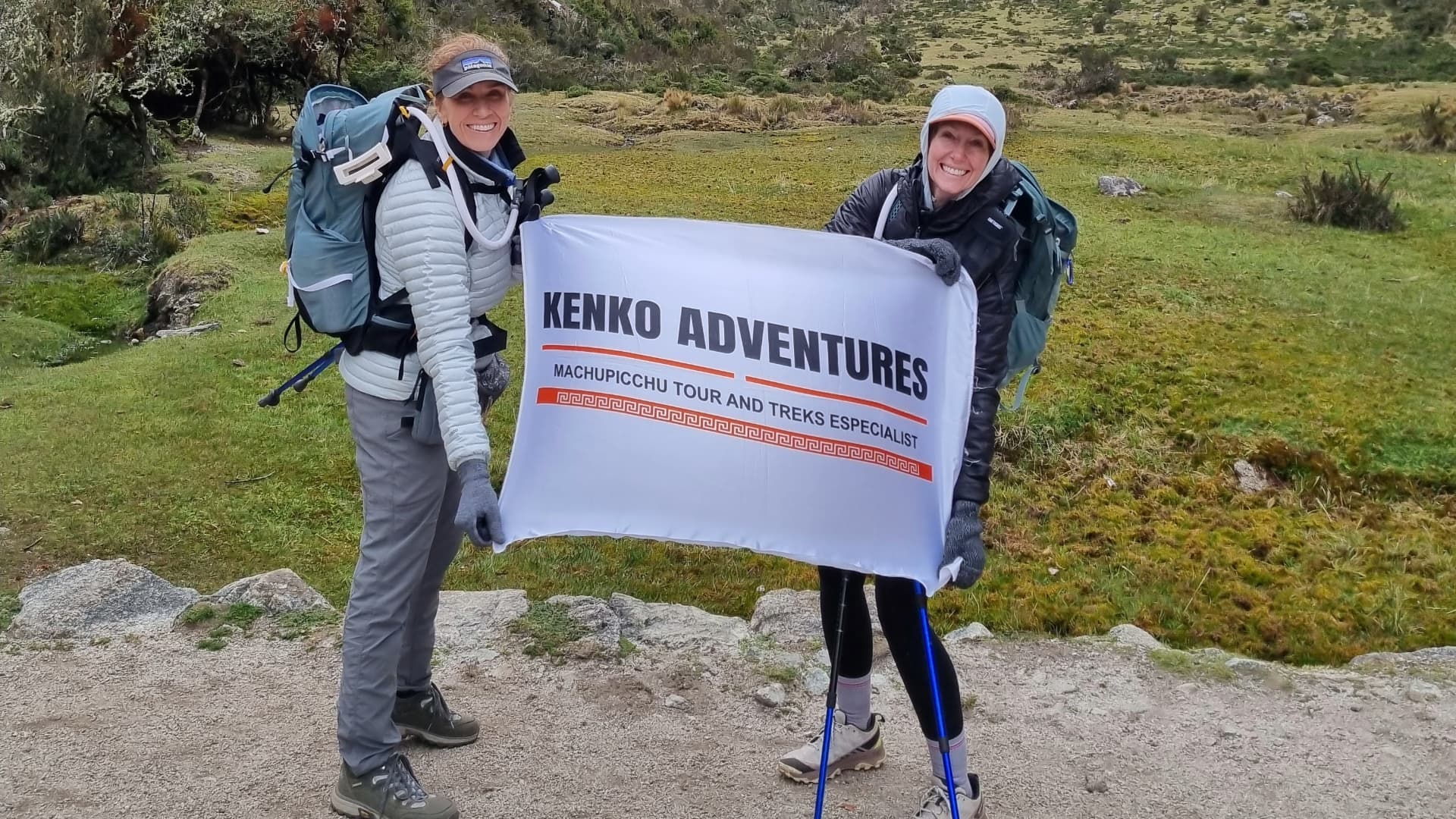 Two women smiling and holding the Kenko Adventures flag, standing against a backdrop of majestic Andean mountains and clear skies. Two women smiling and holding the Kenko Adventures flag, standing against a backdrop of majestic Andean mountains and clear skies.