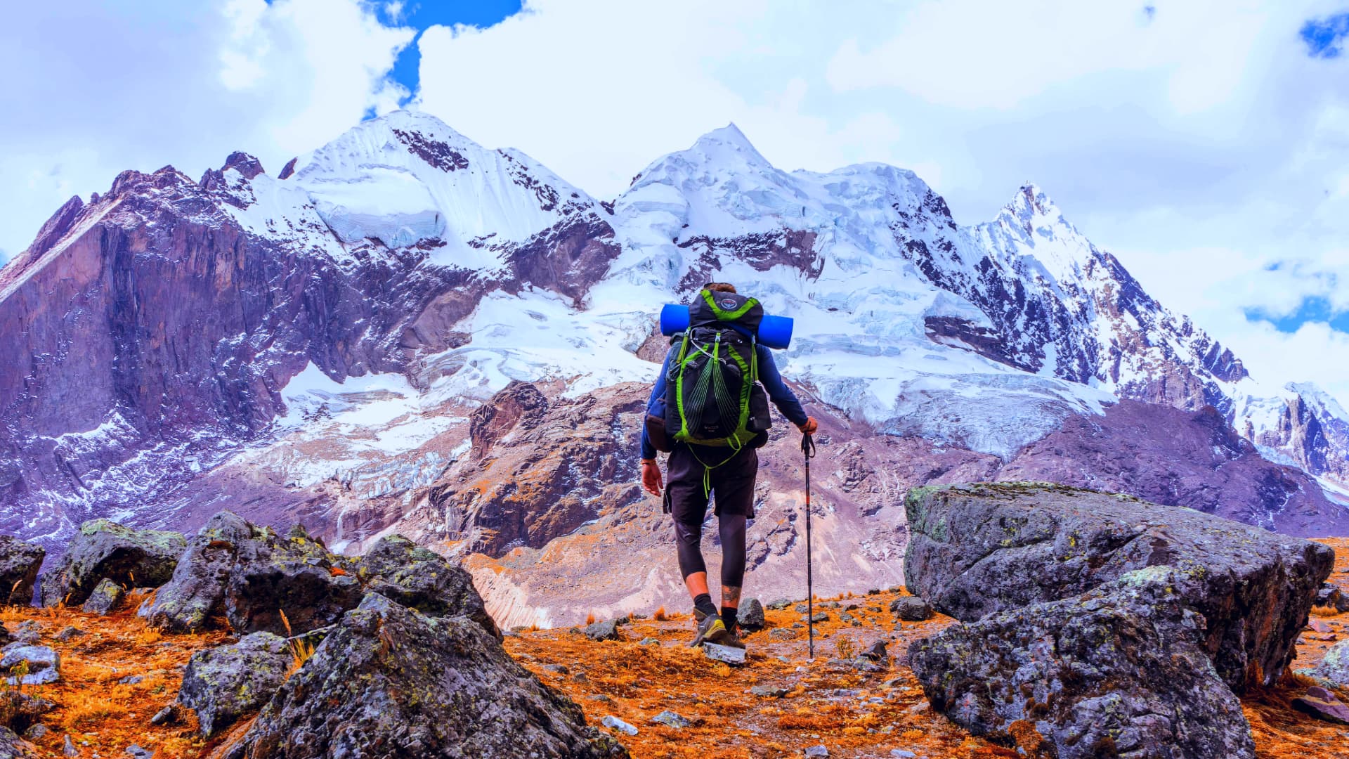 Hiker with a large backpack walking over rocky terrain towards towering snow-covered Andean peaks - Kenko Adventures