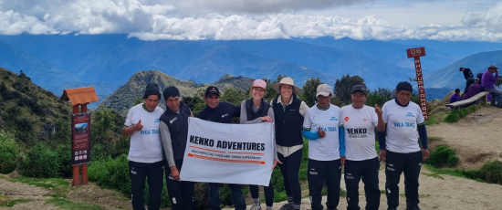 A group of tourists holding a Kenko Adventures flag in the Sacred Valley of the Incas, with terraced hills and towering mountains in the background.