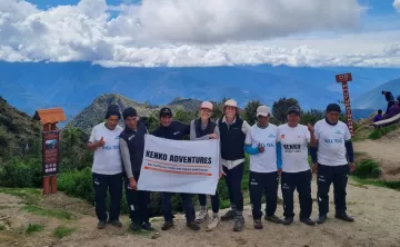 A group of tourists holding a Kenko Adventures flag in the Sacred Valley of the Incas, with terraced hills and towering mountains in the background.