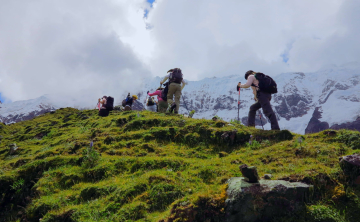 Hikers climbing a steep grassy hill on the Salkantay Trek, surrounded by vibrant greenery and snow-capped peaks in the background under a cloudy sky.
