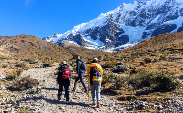 Hikers walking along the rocky trail of the Salkantay Trek, with towering snow-capped peaks of the Peruvian Andes in the background beneath a clear sky.